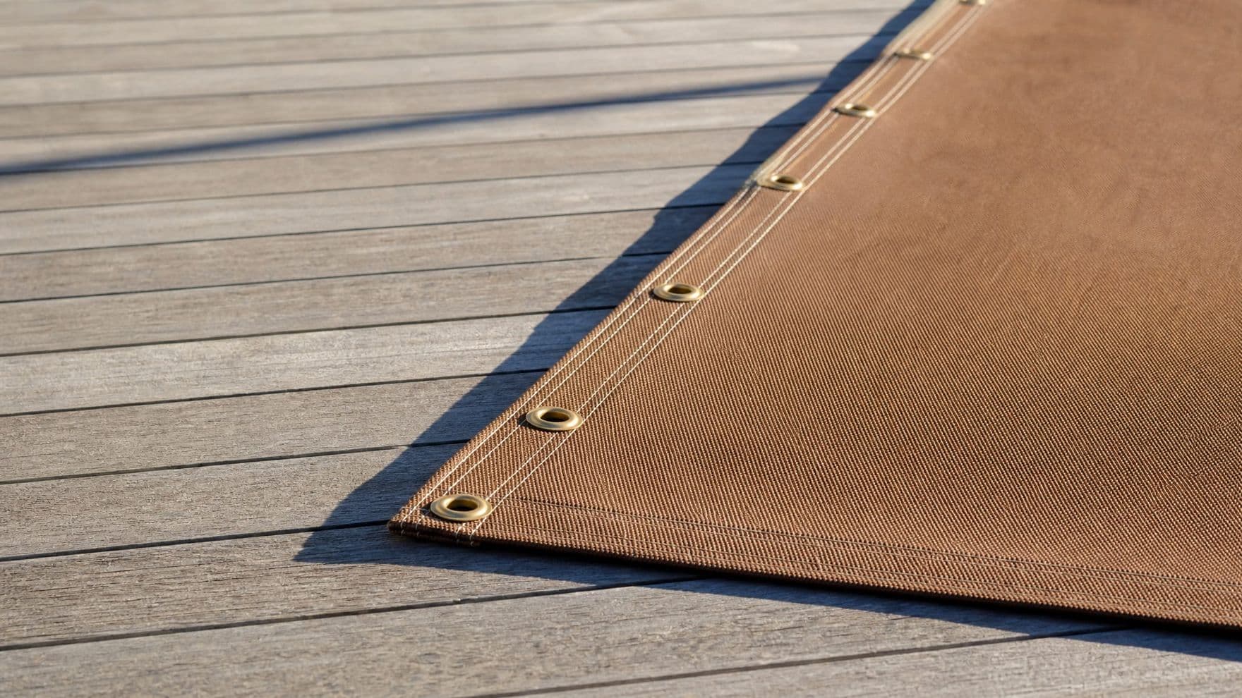 A panel of teak brown acrylic-coated marine canvas lying taut on weathered decking in dry sunlight, its stitched hem and brass eyelets clearly visible.